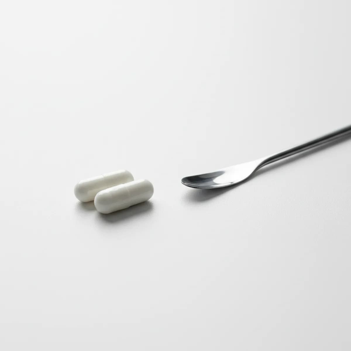 Zwei kleine weiße Kapseln liegen auf einer neutralen weißen Oberfläche neben einem Laborspatel in ruhiger pharmazeutischer Umgebung.